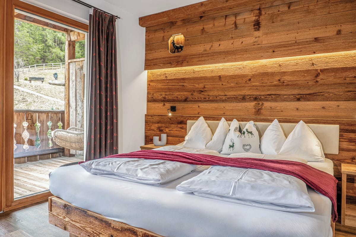 A bedroom with a wooden headboard, a large bed with white bedding and multiple pillows, a nightstand with a lamp, and a sliding glass door leading to an outdoor area with a bench and decorative items.