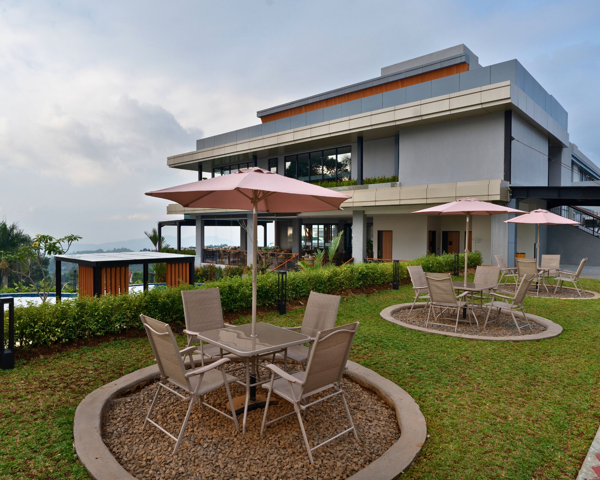 An outdoor seating area with tables and chairs, surrounded by a low hedge and a view of a modern building.