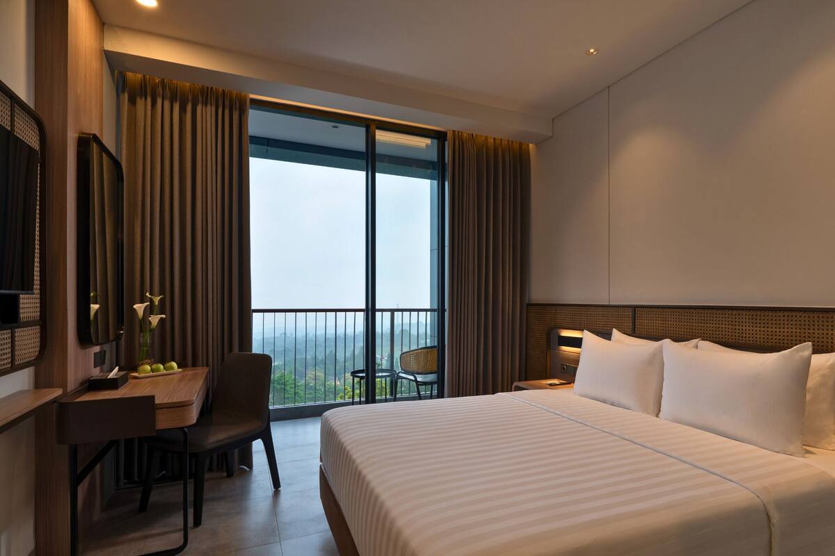 A hotel room with a large bed, a desk, and a chair. There is a balcony with curtains and a view of the city.