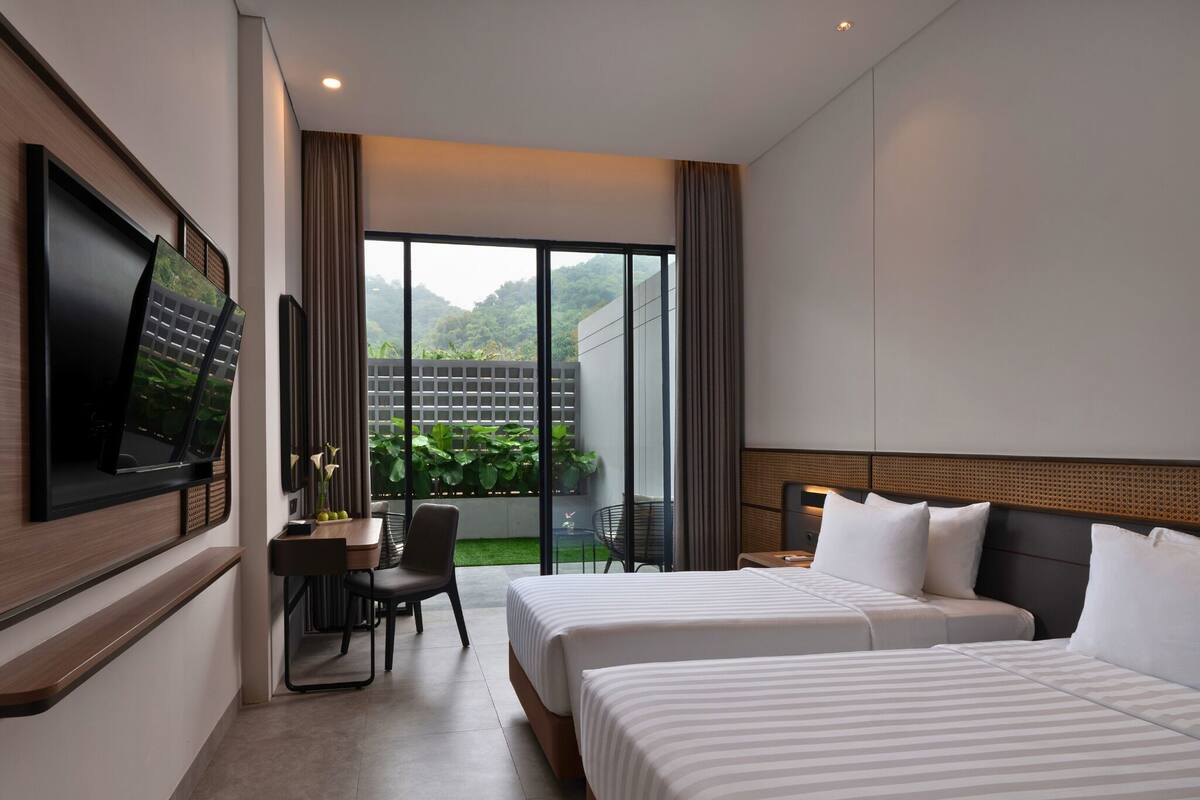 A hotel room with two beds, a desk, a chair, a TV, and a large window with a view of greenery.