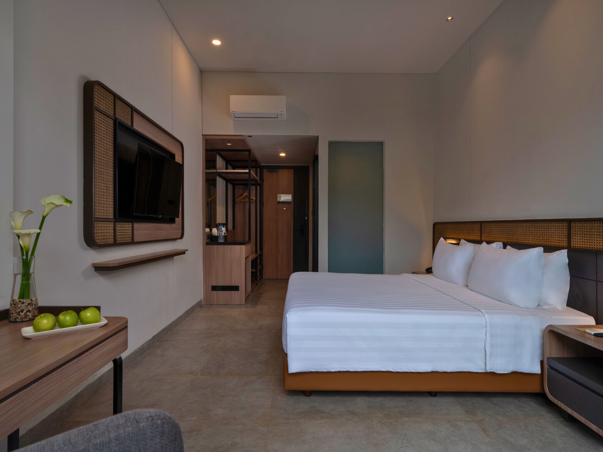 A modern hotel room with a large bed, a flat-screen TV mounted on the wall, a small desk with a vase of flowers, and a kitchenette area.