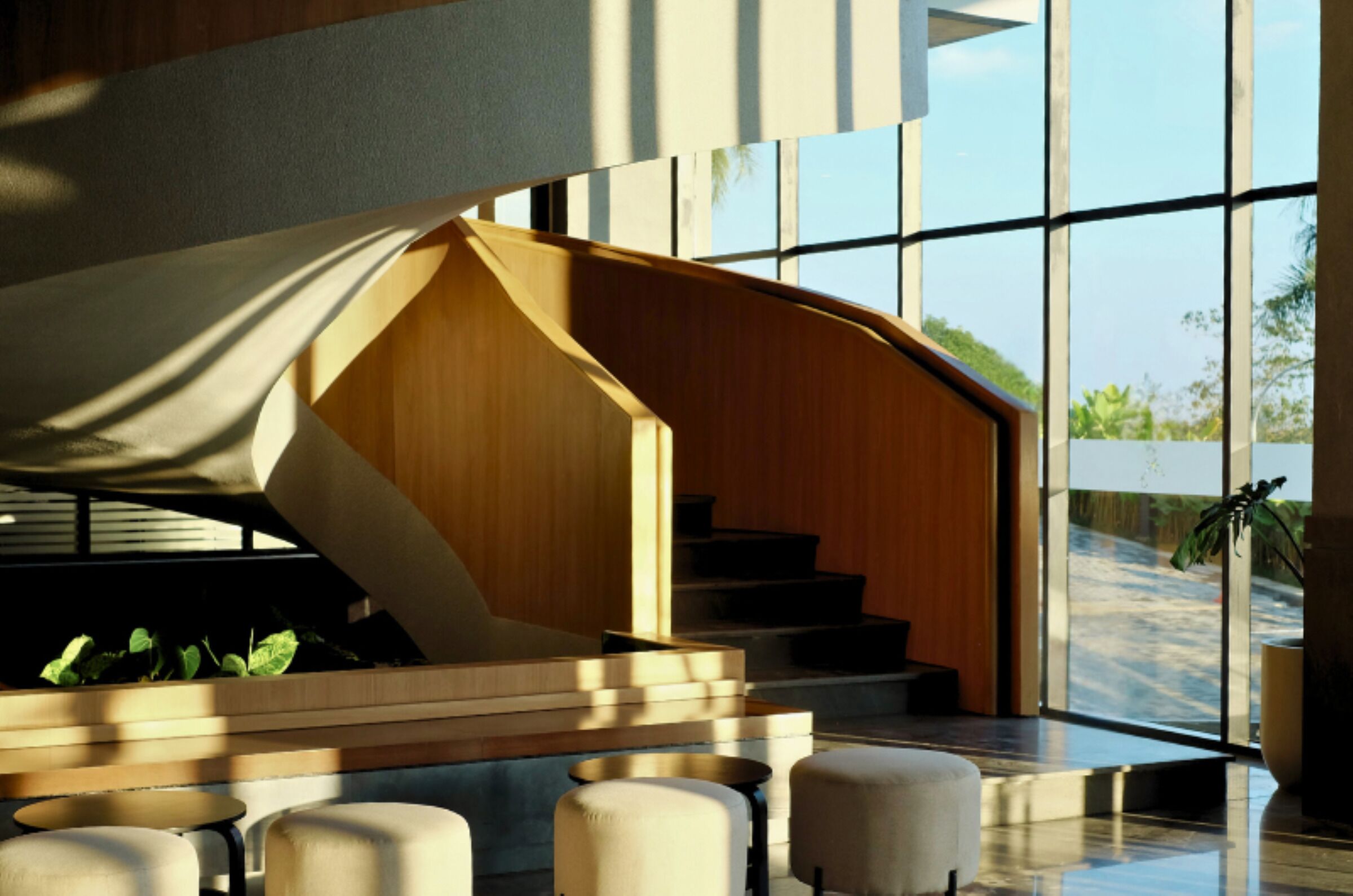 A modern interior with a wooden staircase and seating area, large windows offering a view of greenery outside.