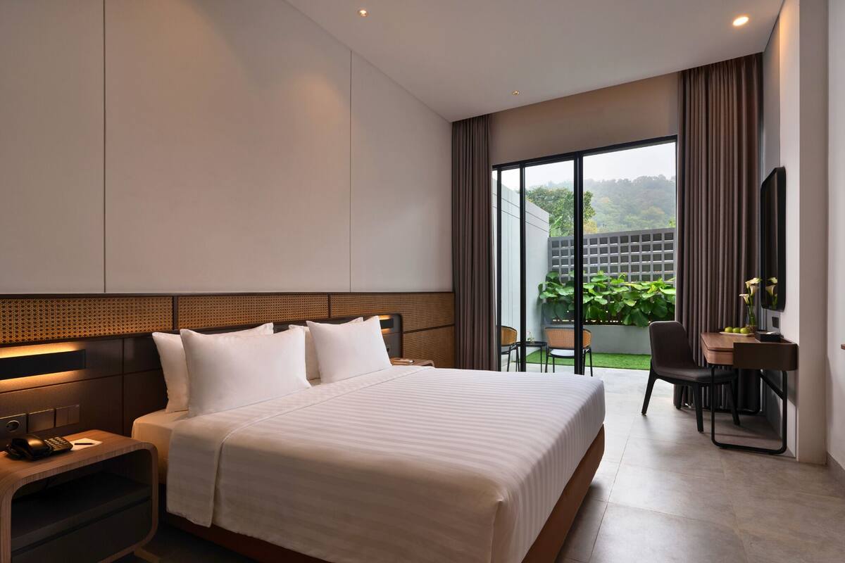 A modern hotel room with a large bed, a desk, and a view of a garden through a sliding door.