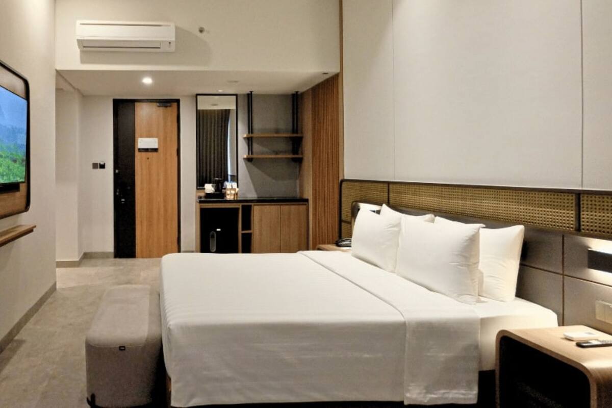 A modern hotel room with a large bed, a bedside table, a small desk, a mirror, and a TV mounted on the wall.