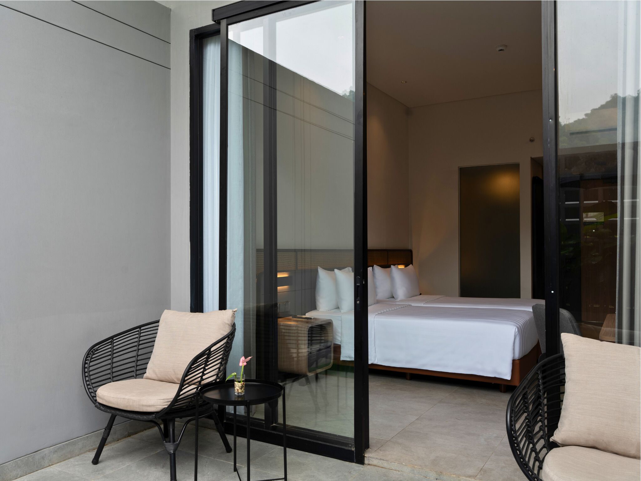 Quarto de hotel moderno com uma cama grande, um espaço de estar pequeno com uma cadeira e mesa, e uma porta de vidro que dá para uma varanda.