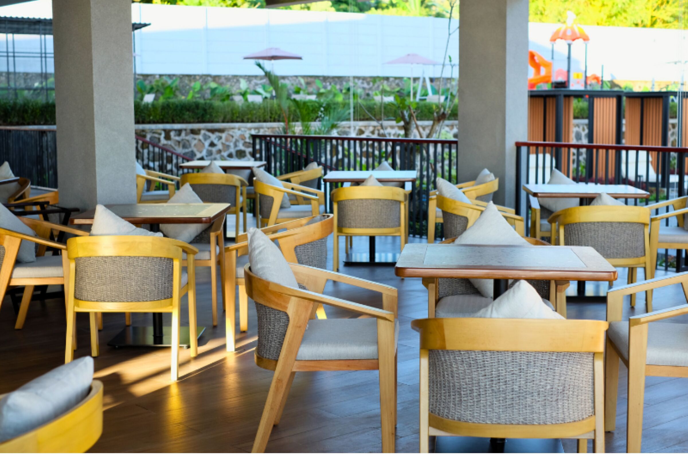 A restaurant with wooden tables and chairs, outdoor seating, and a view of greenery.