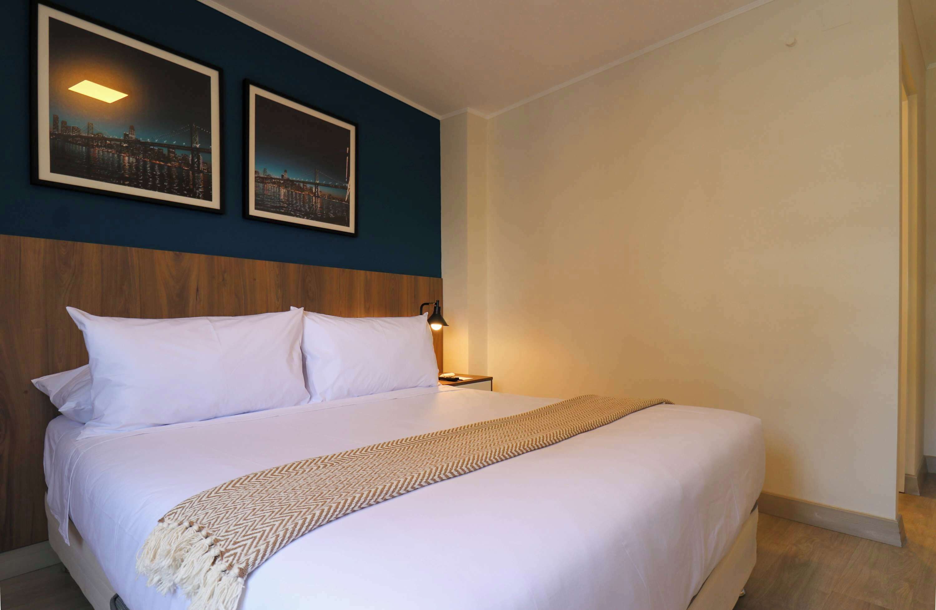 Een net opgemaakt bed met witte lakens en een deken met een patroon, een houten hoofdbord en twee ingelijste foto's aan de muur.
