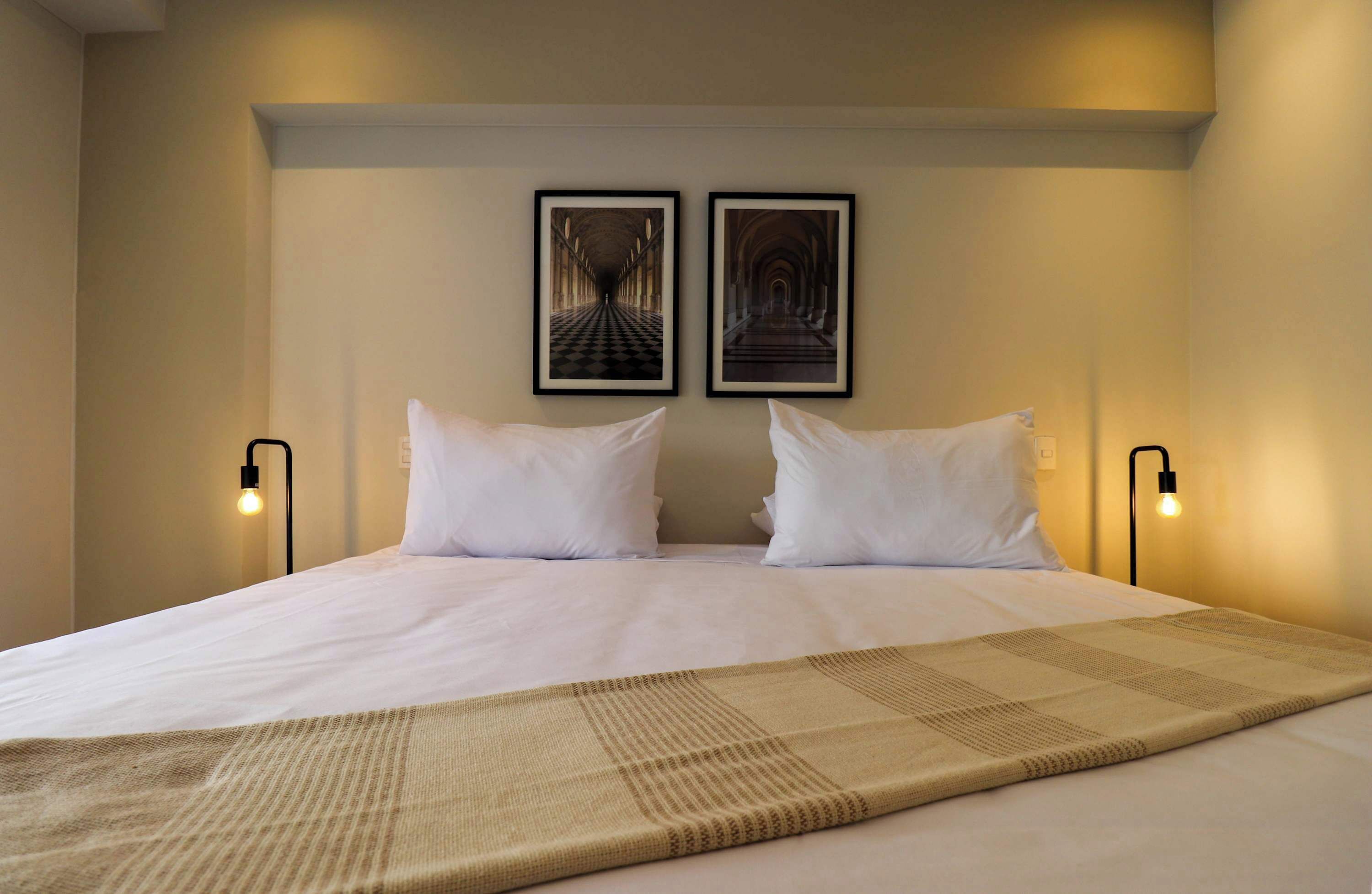 A neatly made bed with white linens and a beige throw blanket, two framed pictures on the wall, and bedside lamps.