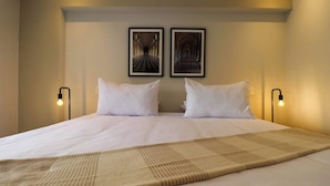 A neatly made bed with white linens and a beige throw blanket, two framed pictures on the wall, and bedside lamps.