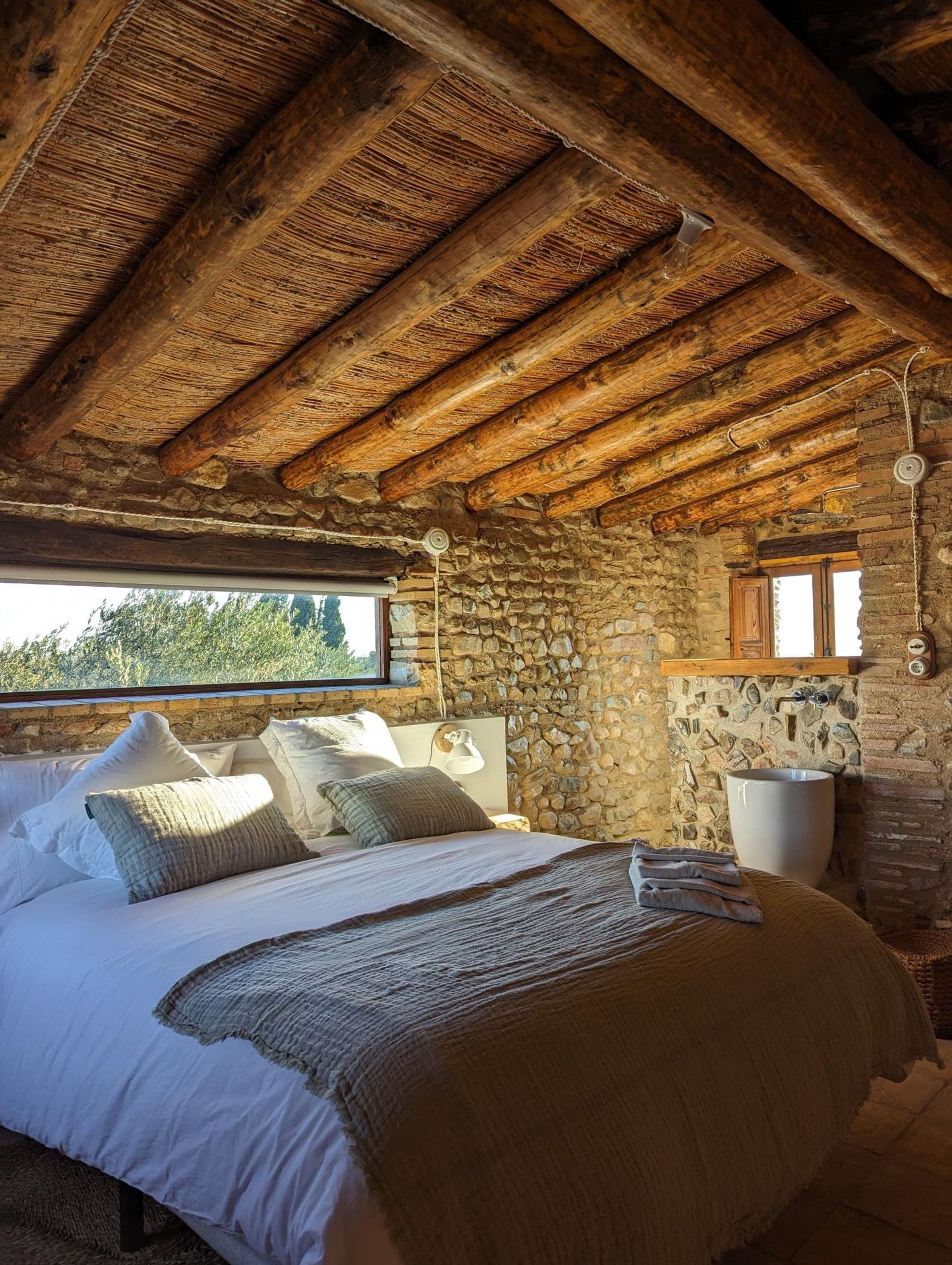 Un dormitorio rústico con pared de piedra, vigas de madera, una cama grande con almohadas, un lavabo de piedra y una ventana con vistas a la vegetación.