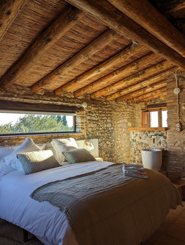 A rustic bedroom with a stone wall, wooden beams, a large bed with pillows, a stone sink, and a window overlooking greenery.
