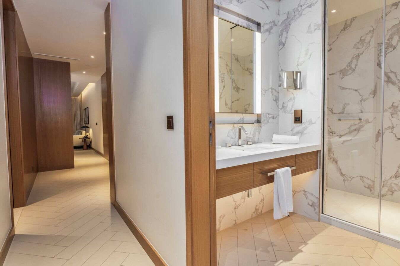 A modern bathroom with a marble vanity, a glass shower enclosure, and a hallway leading to a bedroom.