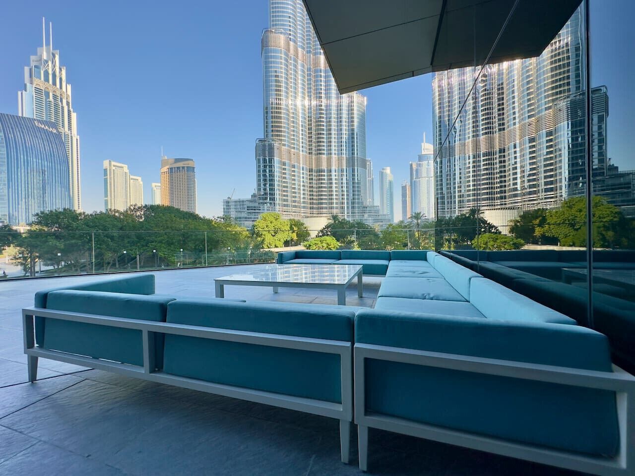 Un salon sur le toit, doté d’une table en verre et de sièges bleus, offrant une vue panoramique sur la ville, avec ses gratte-ciel modernes.