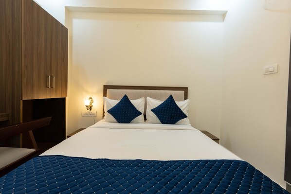 A hotel room with a large bed, two pillows, a bedside table with a lamp, and wooden furniture. - Adhidhani Boutique Hotel (Bengaluru)