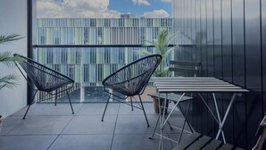 Ein Balkon mit zwei schwarzen Gestellstühlen, einem kleinen Tisch und Blick auf ein modernes Gebäude mit vertikalen Fenstern.