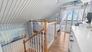 Une chambre avec un escalier en bois, un lit, un tableau représentant des voiliers et une vue sur la mer par la fenêtre.