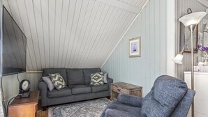 Un salon chaleureux avec un canapé gris, un fauteuil bleu, un tapis à motifs et une petite table en bois sur laquelle se trouve une horloge.