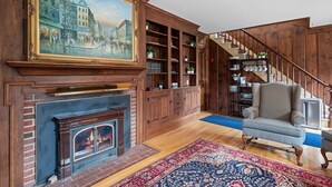 A cozy living room with a fireplace, wooden bookshelves, a large painting, and a staircase.