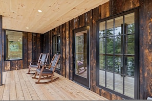 Una terraza de madera con mecedoras y ventanas amplias con vistas a un bosque.