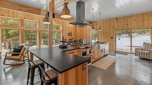 A spacious kitchen with wooden walls and large windows, a central island with bar stools, and a view of a snowy landscape. - Ski Cabin w Hot Tub Gas Fireplace Near Cranmore (Conway)