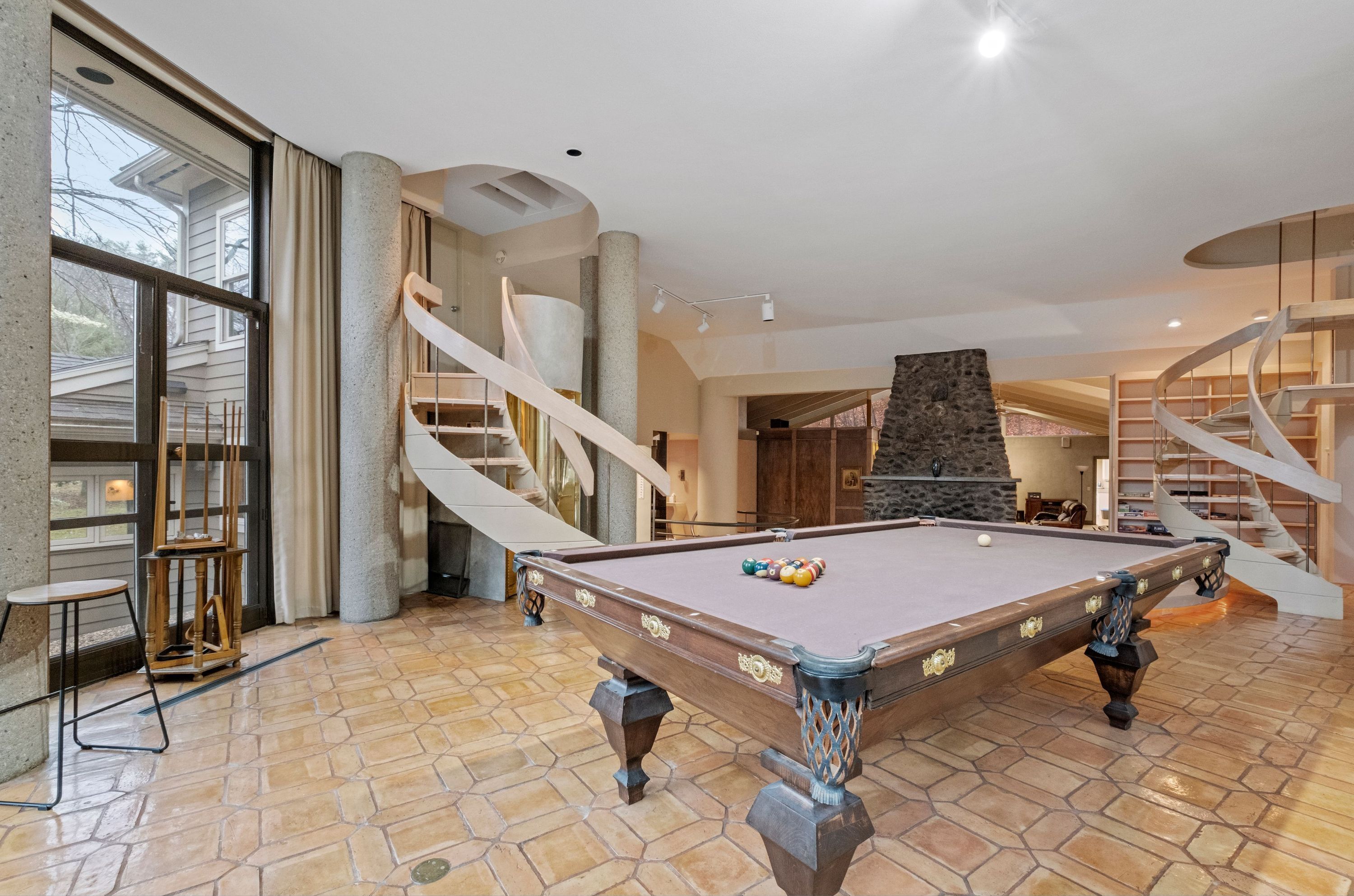 A pool table in a spacious room with a stone floor, a spiral staircase, and a stone fireplace.