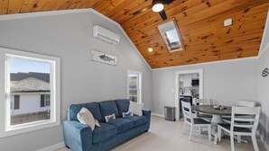Un moderno salón con un sofá azul, una mesa de comedor blanca y sillas. Hay una zona de cocina con armarios blancos y una estufa. La habitación tiene un techo inclinado con vigas de madera y un tragaluz.