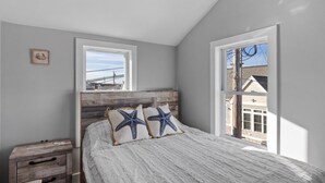 A bedroom with a wooden headboard, a bed with starfish pillows, a nightstand, and a window with a view of buildings. - Modern Condo Perfect For Winter Rental (Hampton)