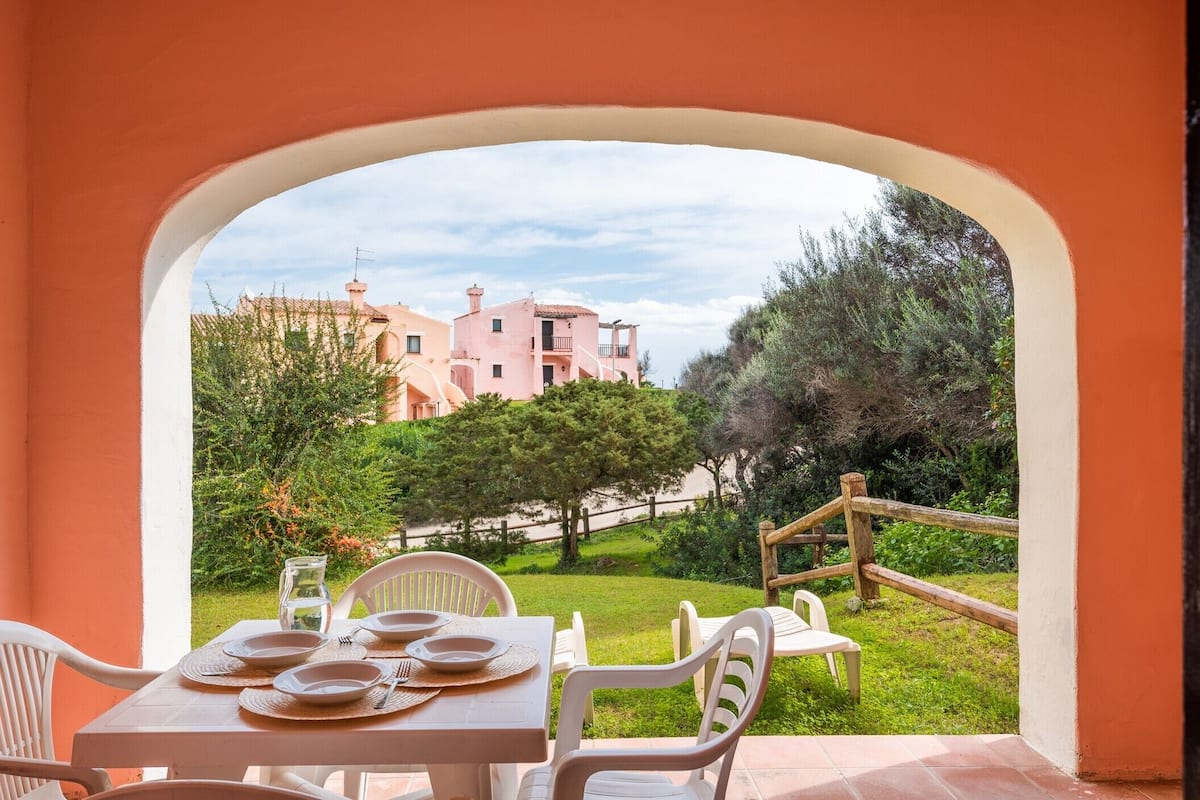 A dining area with a table set for four, overlooking a garden and a pink building with a wooden fence.