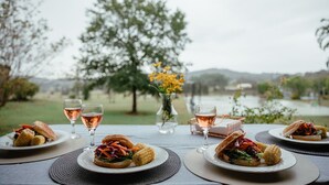A table set with plates of sandwiches, a vase with yellow flowers, and glasses of rosé wine, overlooking a lake.