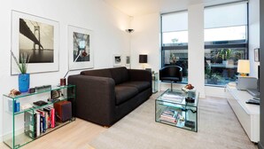 A modern living room with a dark sofa, glass coffee table, and bookshelves. - Zen By The Water, by Veeve (London)