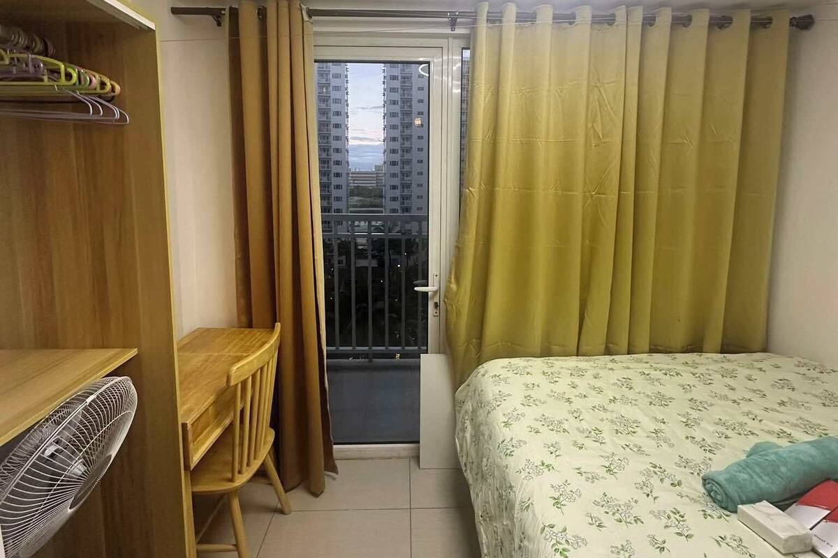 A compact bedroom with a bed, a desk, and a wardrobe. There is a window with curtains, and an air conditioning unit is mounted on the wall.
