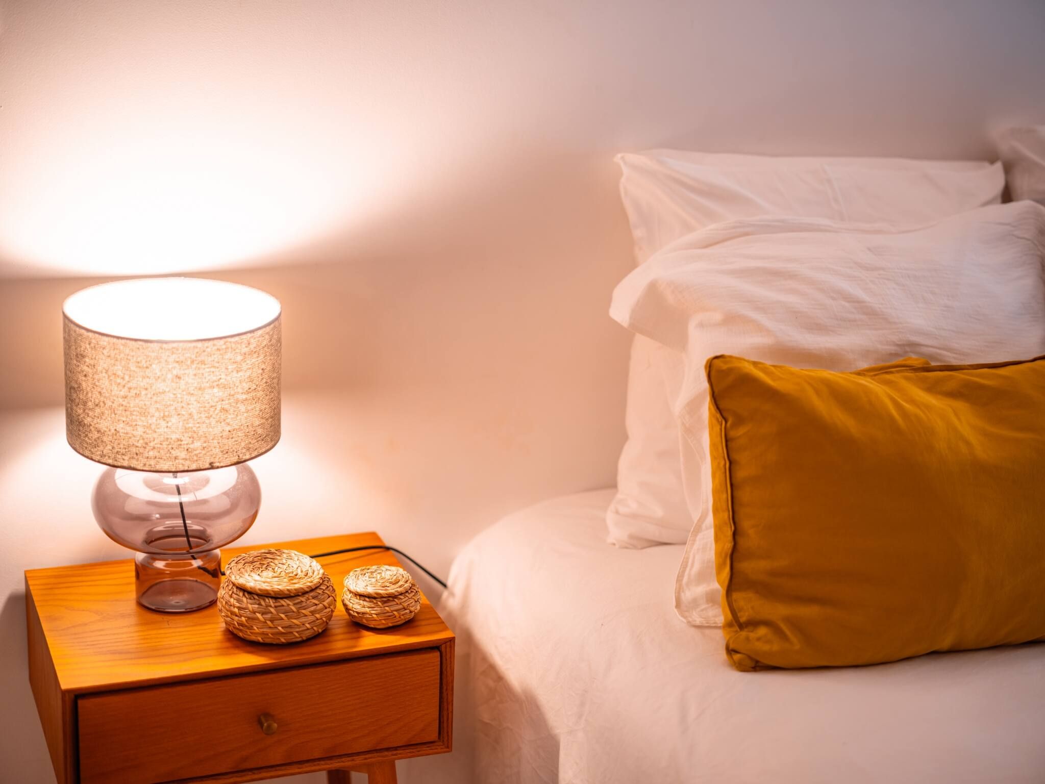 A neatly made bed with white and yellow pillows, a bedside lamp, and two woven baskets on a wooden nightstand.