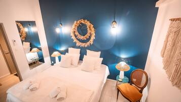 A neatly made bed with white linens and towels, a round woven wall decoration, and a chair with a woven backrest.