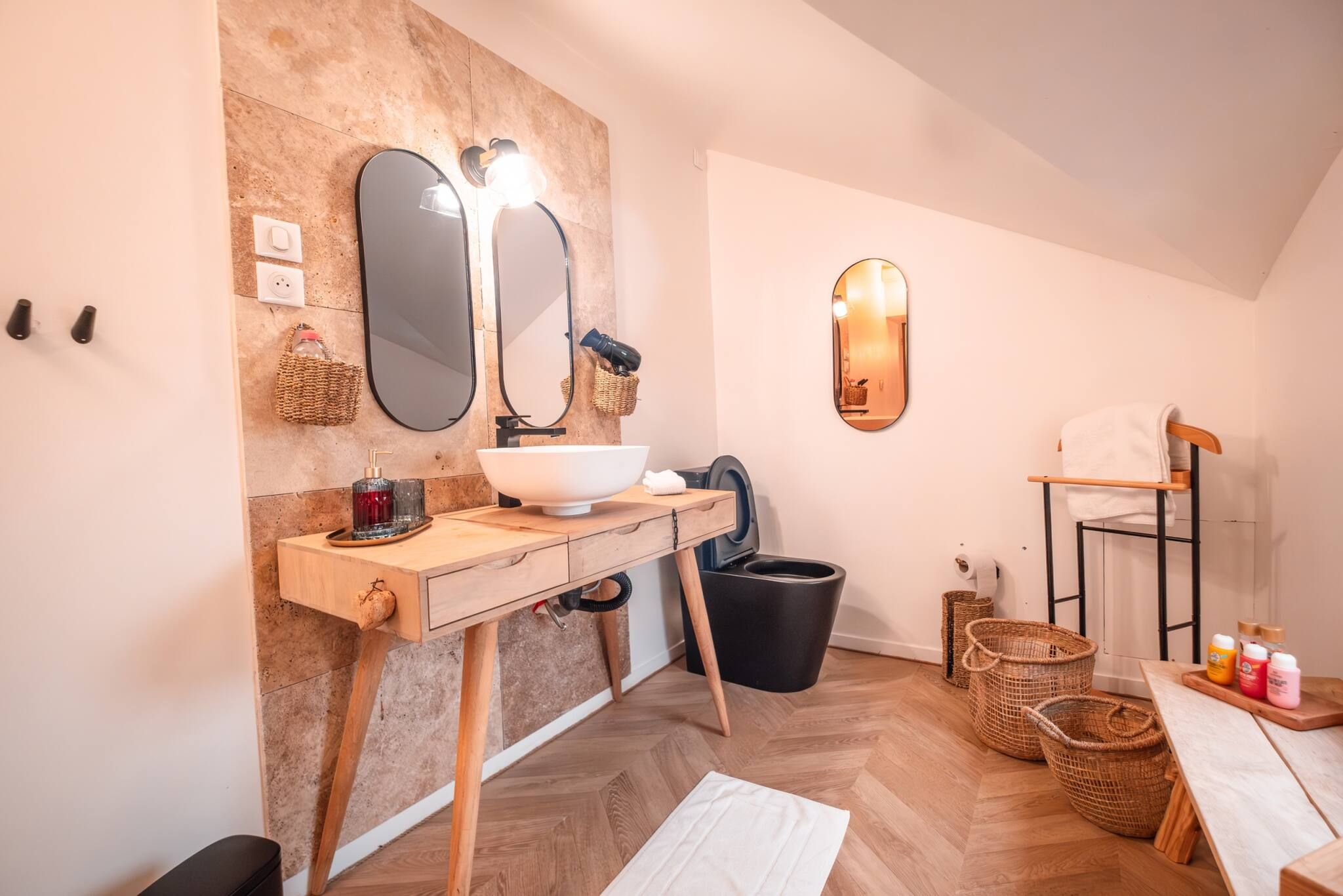 Un baño moderno con tocador de madera, un espejo grande y un lavabo. Hay dos cubos de basura negros y una silla de madera.