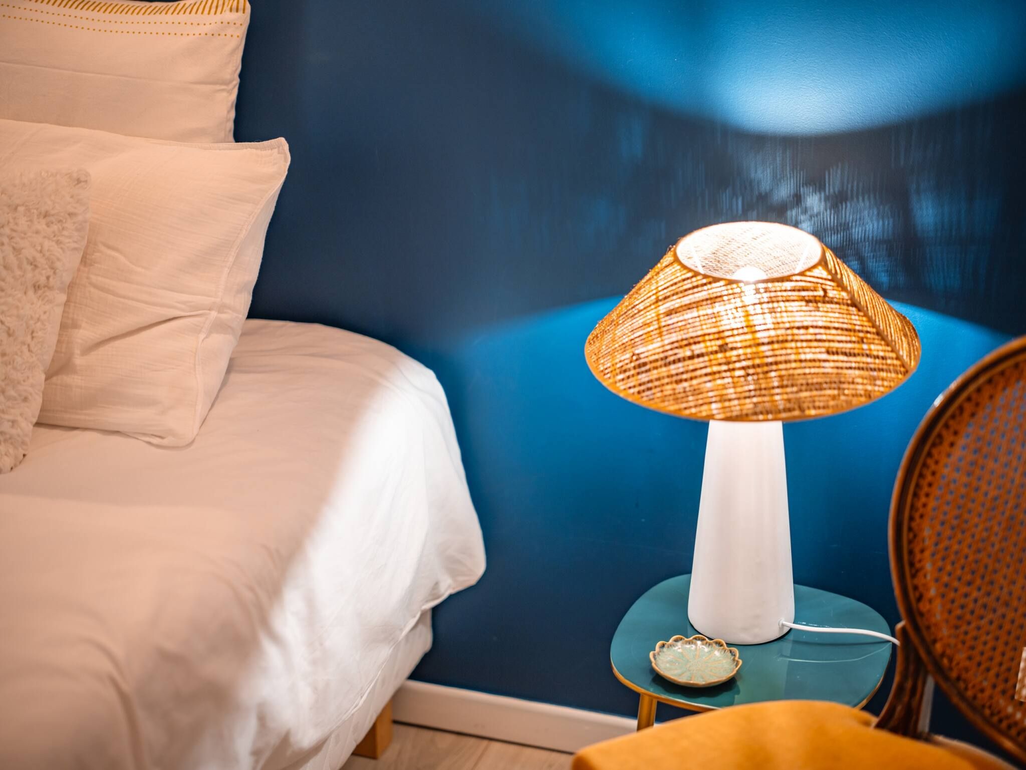 A bed with white linens, a blue wall, a wicker lamp on a small table, and a chair with a woven seat.