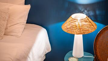 A bed with white linens, a blue wall, a wicker lamp on a small table, and a chair with a woven seat.