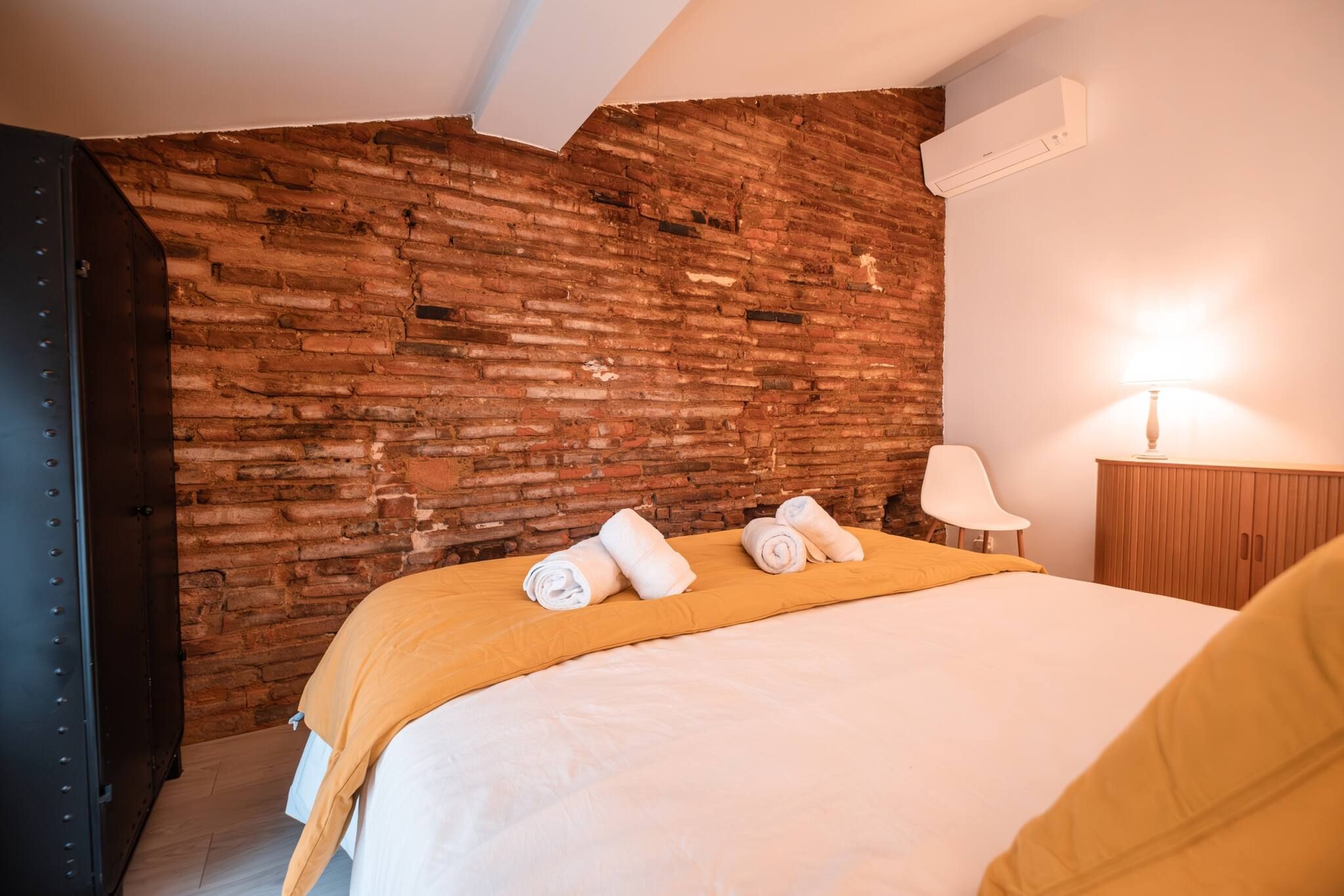 A hotel room with a brick wall, a bed with a yellow bedspread, a white lamp, and a wooden nightstand.