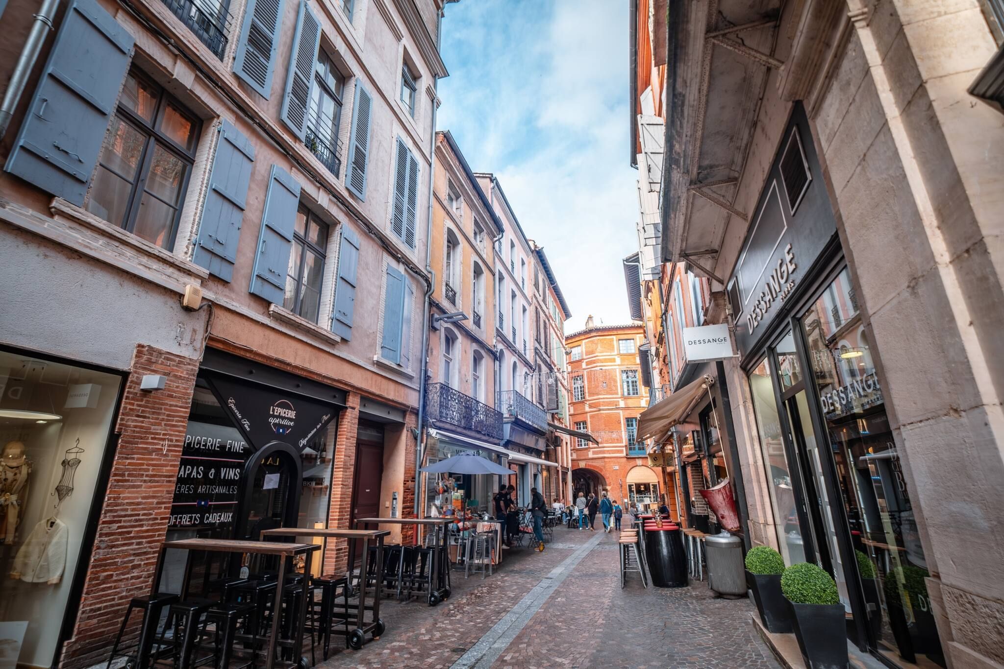 A narrow street with cafes and shops, featuring outdoor seating and brick buildings.