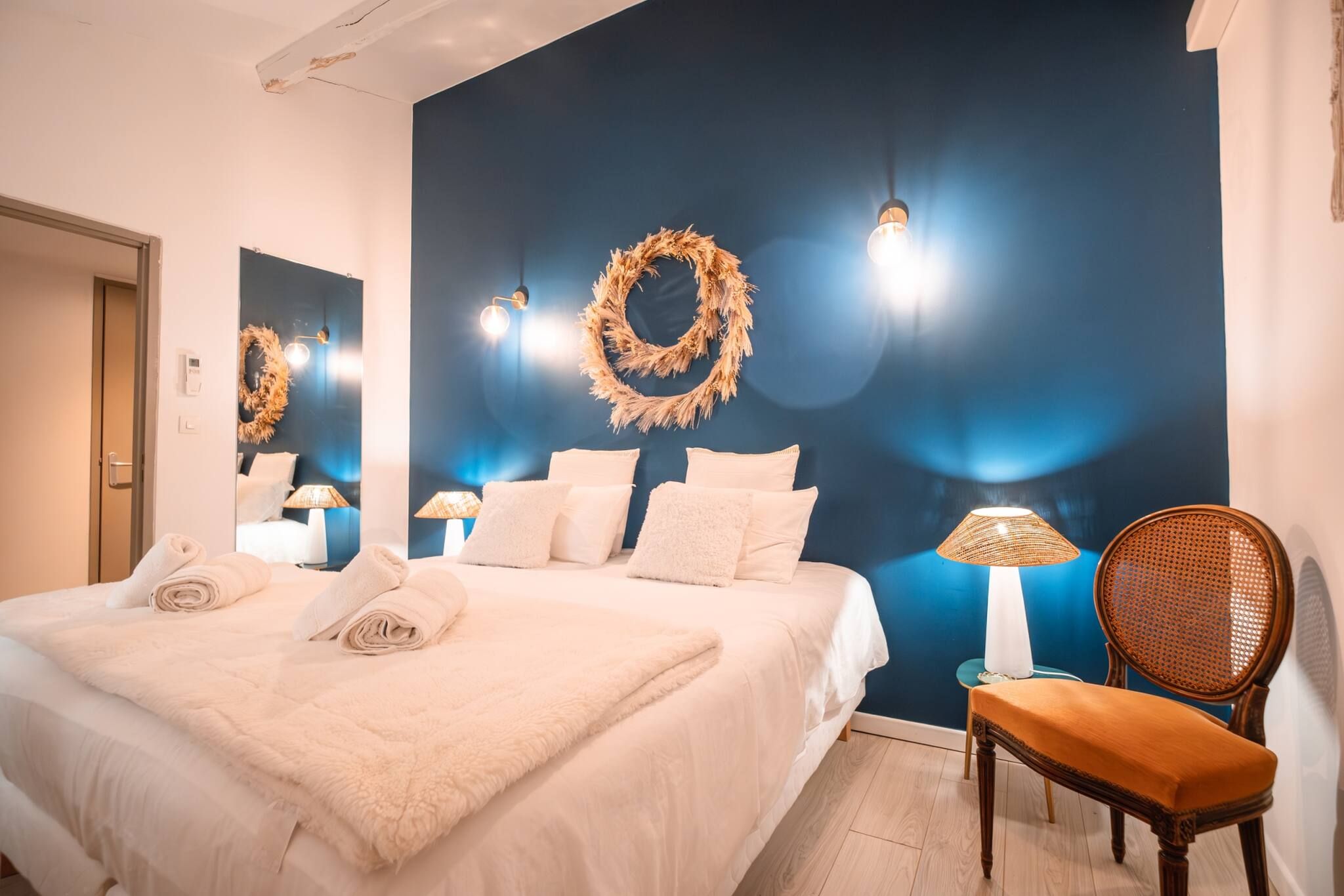 A neatly made bed with a fluffy white blanket, a wooden chair with a cushion, and a wall adorned with a wreath and wall sconces.