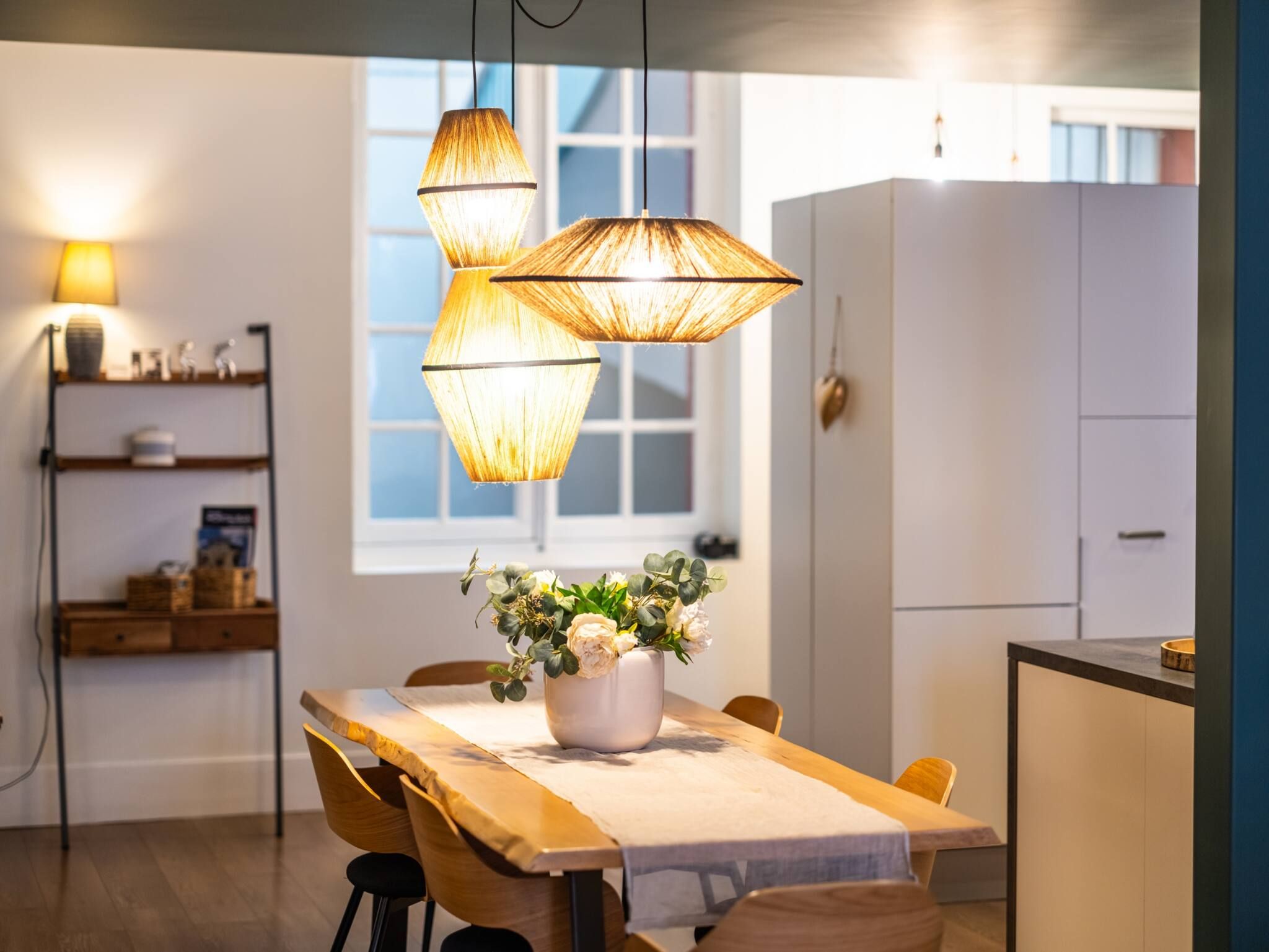 A dining area with a wooden table, chairs, and a vase of flowers. There are hanging lamps above the table.