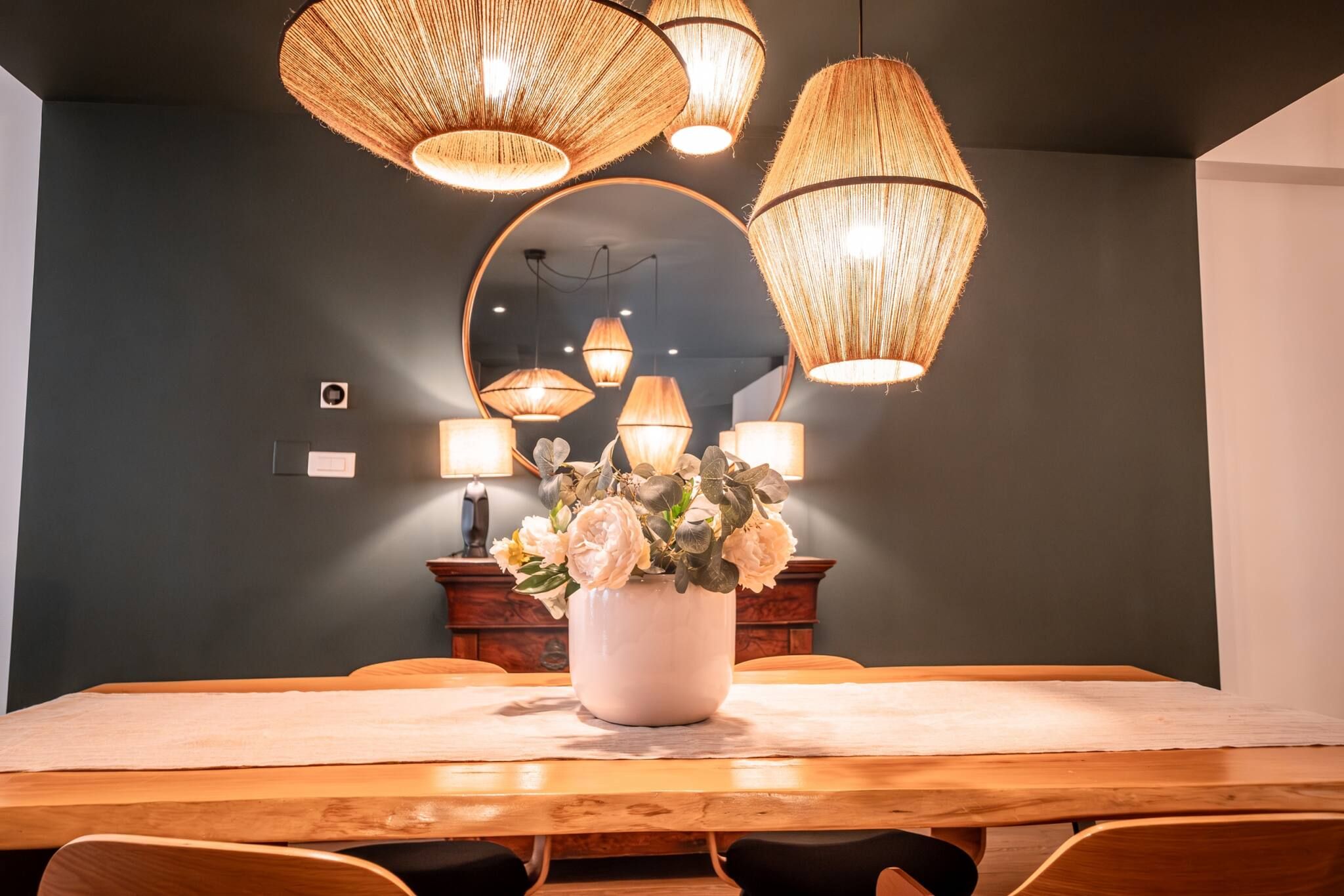 A dining area with a wooden table, white tablecloth, and a vase of flowers. There are wicker lampshades hanging above the table.
