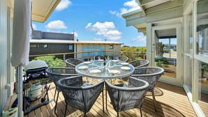 Een overdekt terras met een ronde tafel en rieten stoelen, met uitzicht op een woonwijk met huizen en groen.