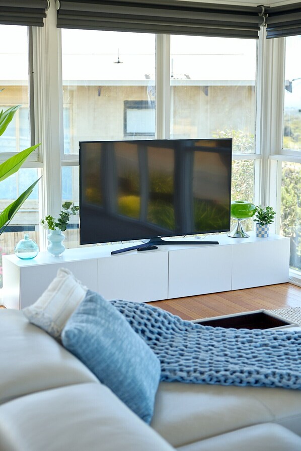 Un salon moderne avec un grand téléviseur à écran plat posé sur un meuble blanc, un canapé agrémenté d’un plaid bleu tricoté et une vue sur un immeuble à travers de grandes fenêtres.