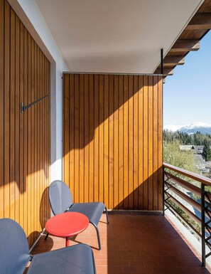 Un balcón con paneles de madera, una mesa roja, dos sillas y vistas a una cadena montañosa nevada.
