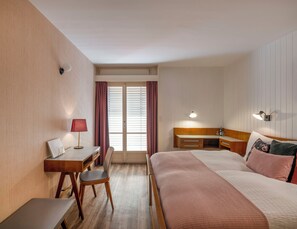 Habitación de hotel con cama, escritorio con silla, mesita de noche, lámpara y ventana con cortinas.