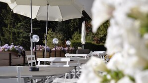 Outdoor tables and chairs with umbrellas, surrounded by flowers and greenery.