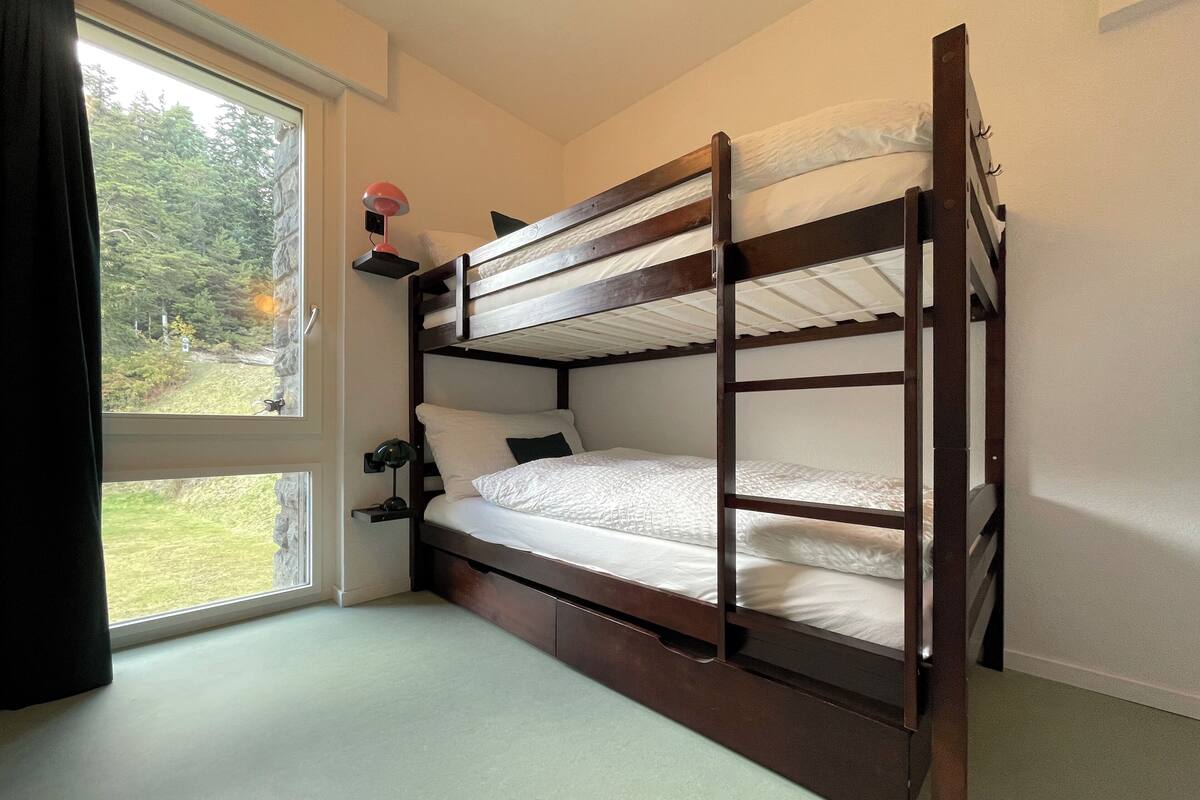 Un lit superposé avec un bureau et une chaise, une fenêtre avec des rideaux et une vue sur la verdure à l’extérieur.
