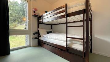 A bunk bed with a desk and chair, a window with curtains, and a view of greenery outside.