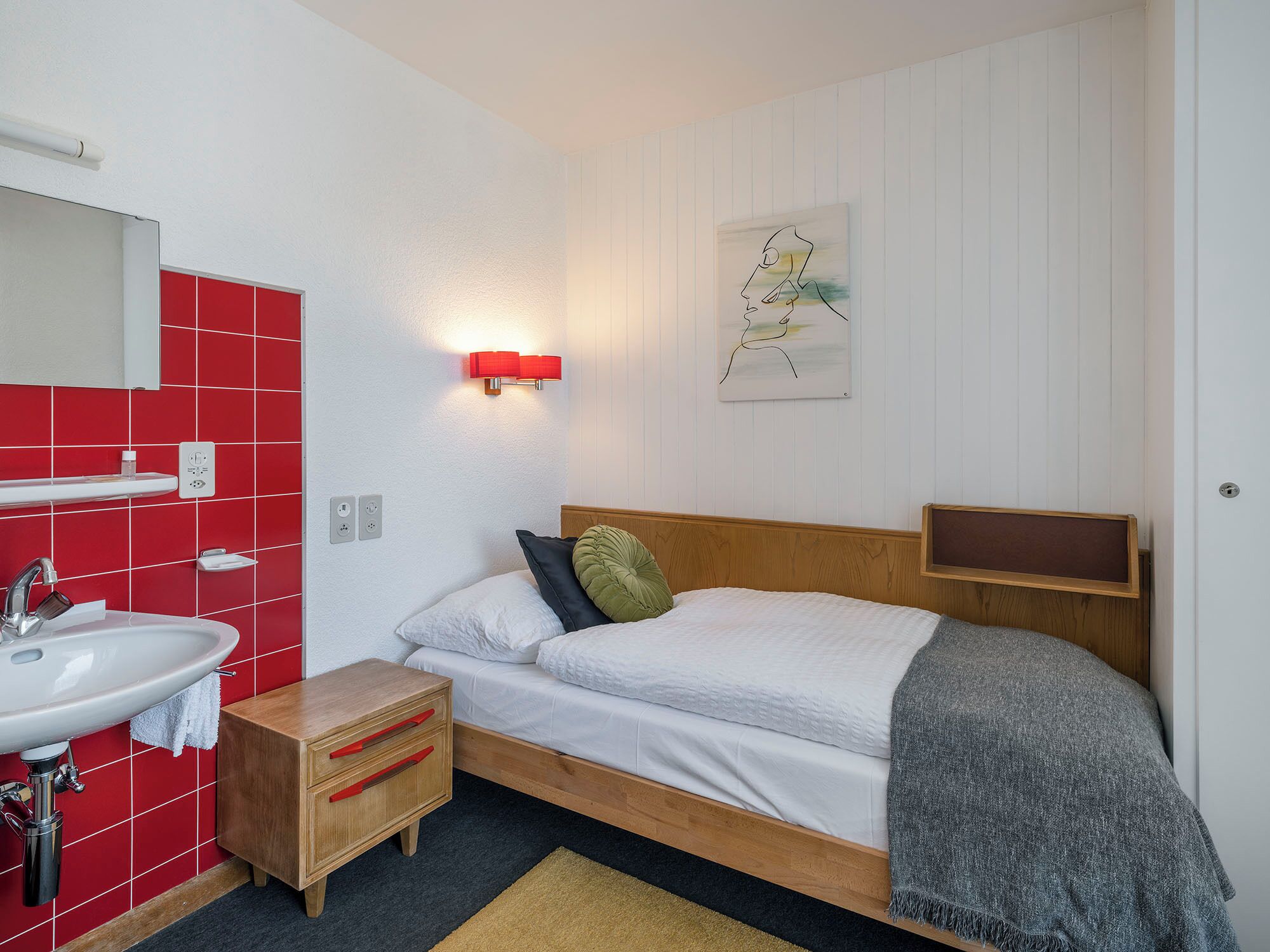 Une chambre avec un lit en bois, du linge de lit blanc, un mur recouvert de carreaux rouges, un lavabo blanc et une petite table de chevet en bois.