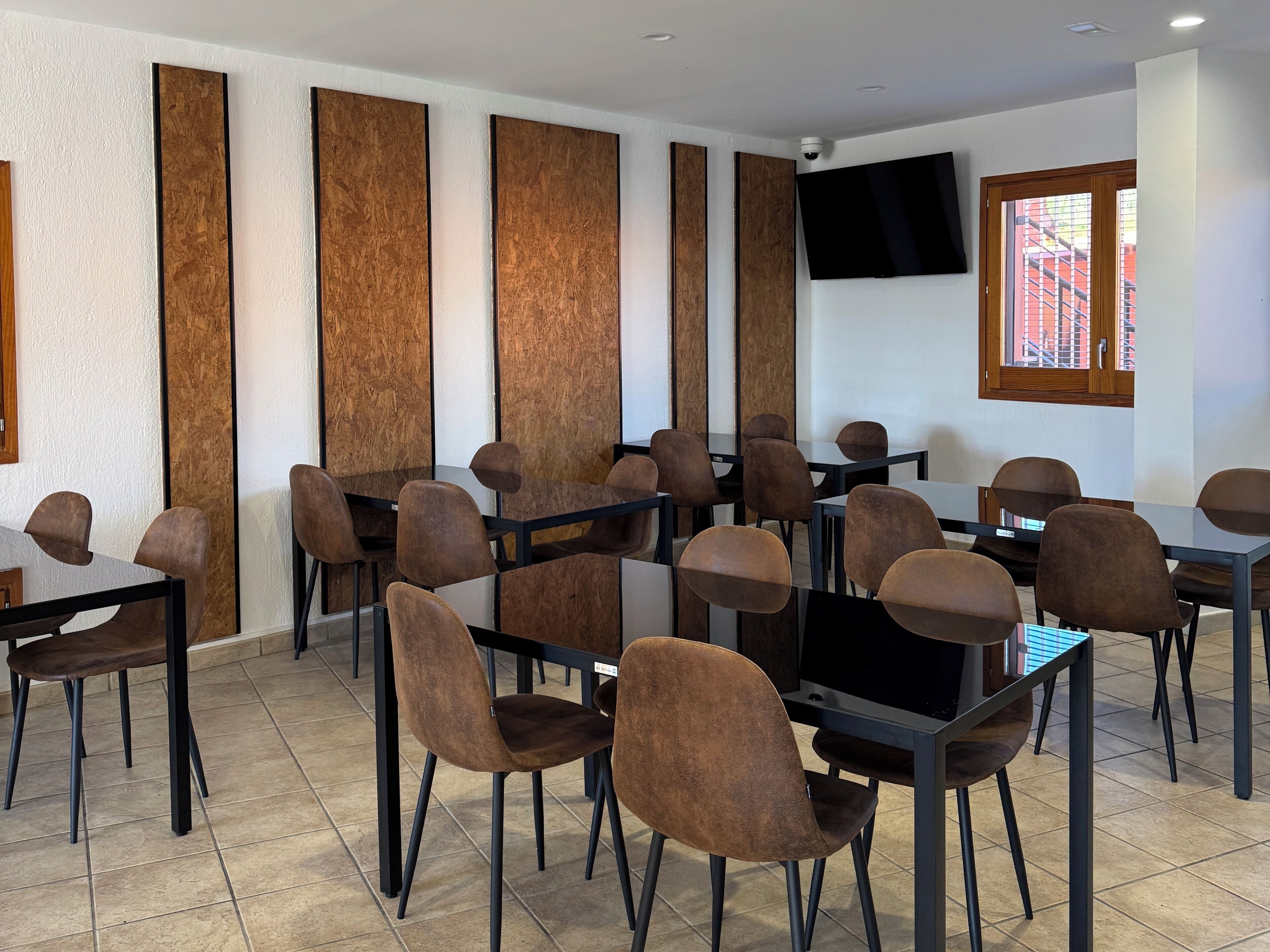 A conference room with multiple tables and chairs, a flat-screen TV mounted on the wall, and a window with a view of buildings outside.